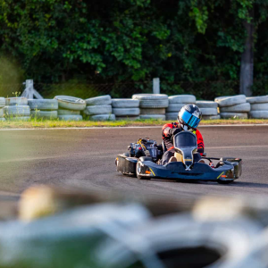 Piloto de kart faz curva em pista de corrida ao ar livre, usando macacão vermelho e capacete com viseira espelhada, com barreiras de pneus e vegetação ao fundo. Esse alt text descreve com clareza o conteúdo da imagem para fins de acessibilidade e também melhora o ranqueamento em mecanismos de busca. Se quiser uma versão mais técnica ou mais informal, posso adaptar.